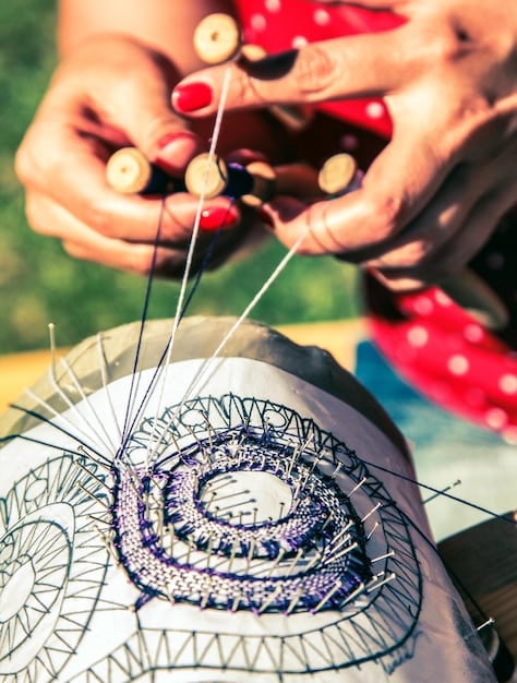 A close-up of a cosplayer's hands meticulously sewing intricate details onto a fabric piece for a costume, surrounded by various sewing tools and design sketches.